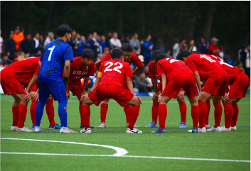 Red soccer players form a huddle on a green pitch before a match, with a blue-clad goalkeeper nearby.